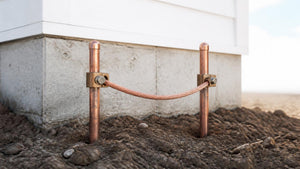 Two copper-bonded ground rods with acorn clamps and bare copper grounding wire at a foundation | Conversions Tech